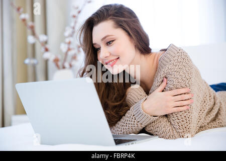 Sorridente piuttosto giovane donna in beige felpa lavorata a maglia giacente sul letto con il computer portatile in camera Foto Stock