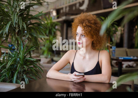 Riflessivo redhead donna con capelli ricci seduti a tavola con lo smartphone nel ristorante e guardando lontano Foto Stock