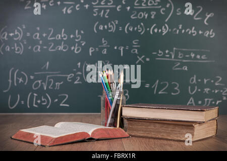 Quaderni e matite giacente su una tavola di legno a banchi di scuola di fronte a una lavagna verde con formule matematiche scuola Foto Stock