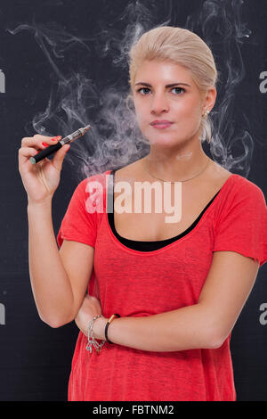 Elegante donna bionda di fumare un e-sigaretta espirando una nube di fumo con gli occhi chiusi in godimento, vista di profilo al buio su un b Foto Stock