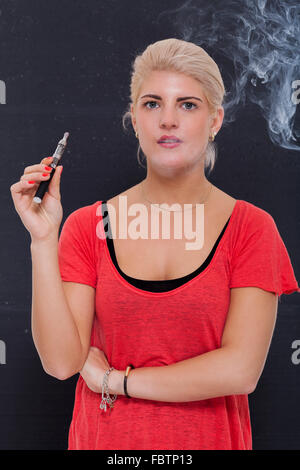 Elegante donna bionda di fumare un e-sigaretta espirando una nube di fumo con gli occhi chiusi in godimento, vista di profilo al buio su un b Foto Stock