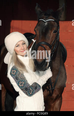 Bella ragazza bionda avvolgente cavallo marrone in inverno agriturismo Foto Stock