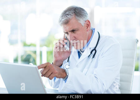 Medico serio lavoro su laptop e avente chiamata telefonica Foto Stock