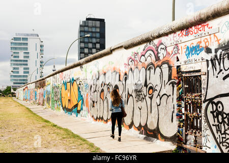 Giovane donna camminare passato graffiti sul muro di Berlino, Muhlenstrasse, quartiere Friedrichshain di Berlino, Germania Foto Stock