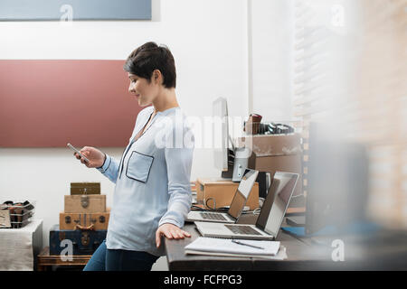 Una donna in un ufficio a casa con una scrivania con due computer portatili, controllando il suo smart phone. Foto Stock