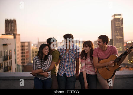 Un piccolo gruppo di amici riuniti su un tetto a terrazza che si affaccia su una città al crepuscolo. Foto Stock