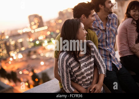 Un piccolo gruppo di amici riuniti su un tetto a terrazza che si affaccia su una città al crepuscolo. Foto Stock