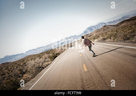 Un giovane uomo che cavalca verso il basso una strada asfaltata nel deserto su uno skateboard. Foto Stock