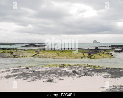Kicker Rock lontano all'orizzonte contro un cielo nuvoloso da una spiaggia di sabbia nera con rocce laviche coperte di verde brillante mossy Foto Stock