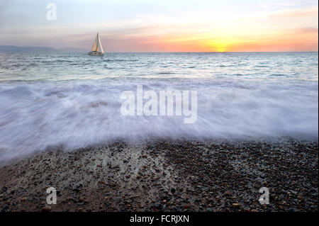 Barca a vela al tramonto è una barca a vela in movimento lungo l'acqua con un oceano frangi onda in primo piano. Foto Stock