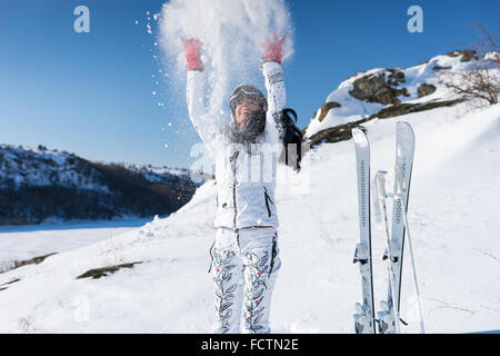 Eccitato Singolo sciatore femmina in bianco doposci e casco gettando la neve sopra la sua testa come essa si erge accanto al montante di sci con collina dietro di lei. Foto Stock