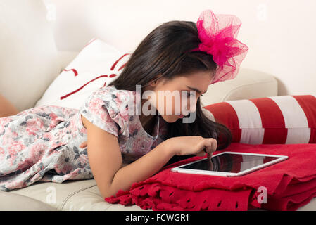 Ragazza giovane navigare su internet su un tablet come lei giace sul suo letto lavorando su un colorato di rosso coperta con un corrispondente nastro in h Foto Stock