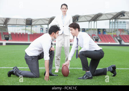 Due uomini di affari inginocchiato sul campo di erba con una palla da rugby con un business woman standing tra di essi Foto Stock