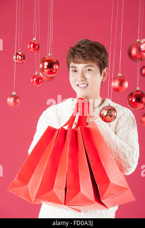 Giovane adulto uomo in bianco maglione rosso di contenimento delle borse per lo shopping sotto la decorazione di Natale e lo sguardo in avanti con un sorriso Foto Stock