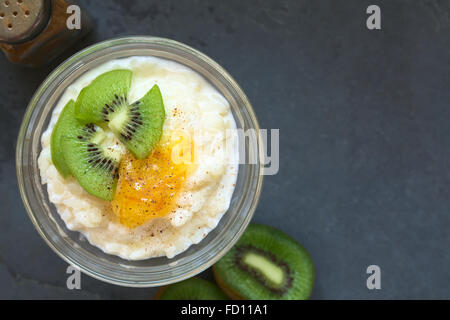 Budino di riso con pezzi di kiwi, marmellata di arancio e cannella in piccola ciotola di vetro, fotografato overhead su ardesia con luce naturale Foto Stock
