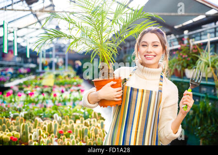 Felice donna attraente giardiniere nel grembiule azienda piccola palma nella pentola e la forcella per il trapianto di piante in serra Foto Stock