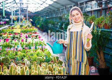 Felice affascinante giovane donna giardiniere nel grembiule a strisce e permanente tenendo garden pala e blue annaffiatoio in serra Foto Stock