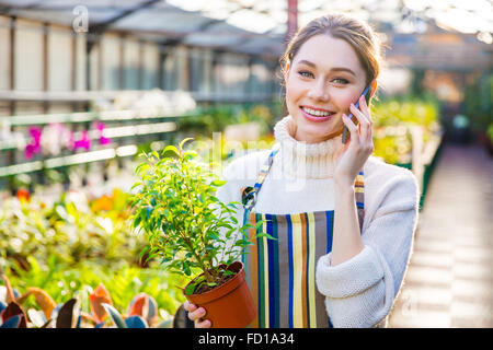 Bella sorridente giovane donna giardiniere nel grembiule a strisce parlando al telefono cellulare e azienda piccola pianta in vaso in aranciera Foto Stock