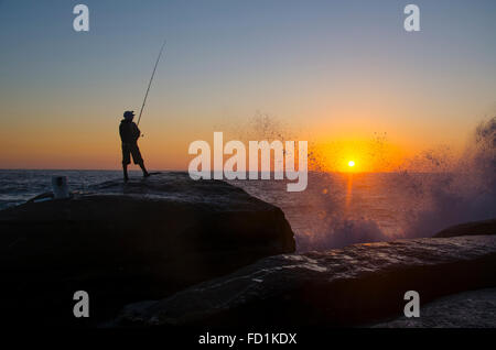 Un pescatore solista si erge, in una posizione che si staglia contro l'alba su una grande roccia, pescando mentre le onde si infrangono contro le rocce sottostanti a Sydney, Australia Foto Stock