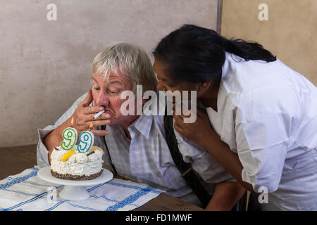 Naughty anziano pensionato lighting la sua sigaretta sulla sua torta di compleanno Foto Stock