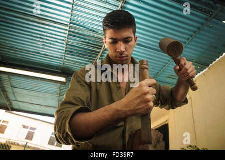 Giovane uomo al lavoro artigiano di apprendimento professione, lavorando con martello e scalpello. L'artista scolpisce un blocco grezzo di legno per fare un Foto Stock