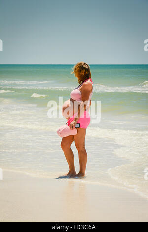 Una vista laterale di una donna in stato di gravidanza, vestito in rosa esponendo il suo bambino bump mentre trampolieri nel surf in spiaggia Foto Stock