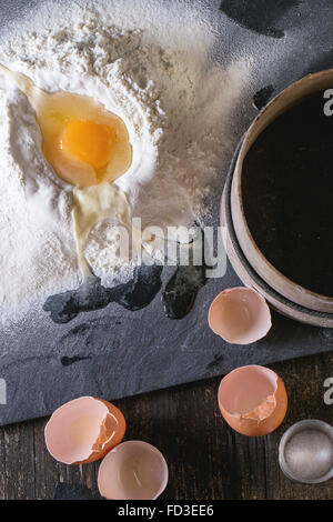 Ingredienti per la pasta facendo. La farina setacciata e brocken uovo su nero ardesia scheda sopra il vecchio tavolo in legno. Stile rustico. Vista superiore Foto Stock