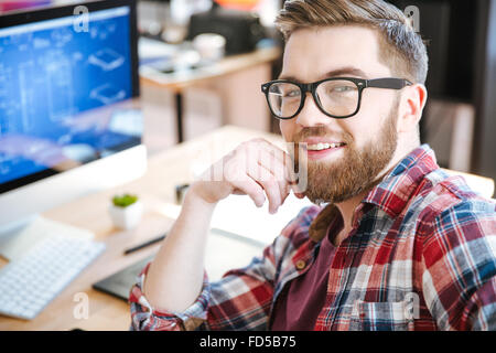 Felice attraente giovane uomo con la barba in bicchieri di lavoro e di progettazione di progetto sul suo computer Foto Stock