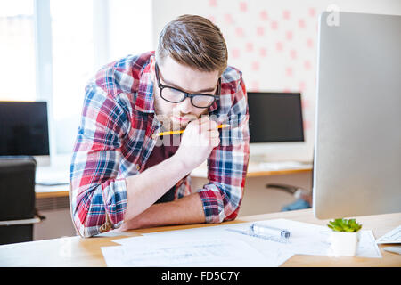 Riflessivo uomo creativo con la barba in bicchieri progettazione di progetto e fare blueprint sul suo luogo di lavoro Foto Stock