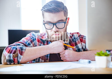 Ritratto di pensieroso barbuto giovane ingegnere in bicchieri di sedere a tavola e creare blueprint Foto Stock