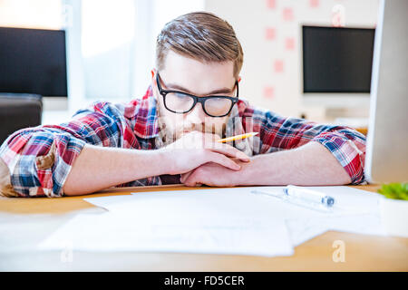 Riflessivo barbuto giovane uomo in bicchieri lavora con blueprits utilizzando una matita e righello in ufficio Foto Stock
