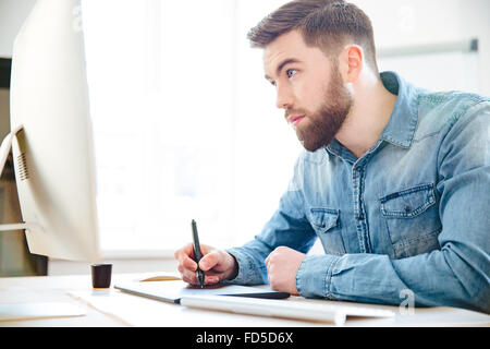 Concentrati bel giovane designer con la barba in maglietta blu disegno usando il computer e tavoletta grafica in ufficio Foto Stock