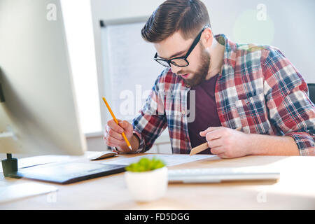 Bello concentrato giovane uomo con la barba lavora con blueprint con un righello e matita in ufficio Foto Stock