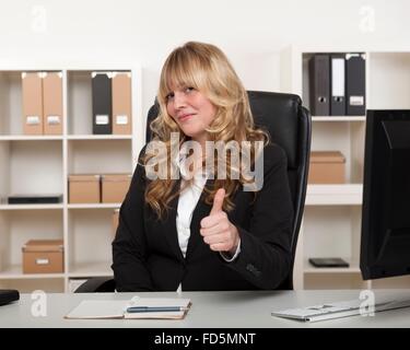 Attraente imprenditrice con ricci lunghi capelli biondi seduto alla sua scrivania in ufficio dando un pollice in alto gesto di approvazione Foto Stock