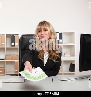 Bella giovane imprenditrice tenendo fuori un pugno di banconote come ella si estende in tutta la sua scrivania concettuale del successo Foto Stock