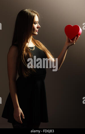 La felicità, il giorno di San Valentino e il concetto di amore. Bella elegante pensieroso donna in abito nero azienda cuore rosso a forma di scatola regalo. Foto Stock