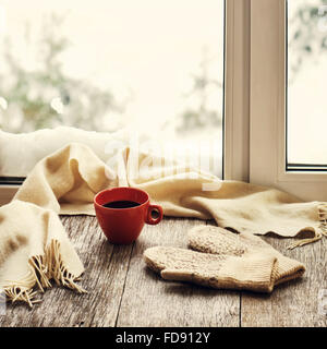 Red tazza di caffè o di tè, beige sciarpa e womans guanti sono situati sul legno stilizzata davanzale. Inverno concetto di comfort di un Foto Stock