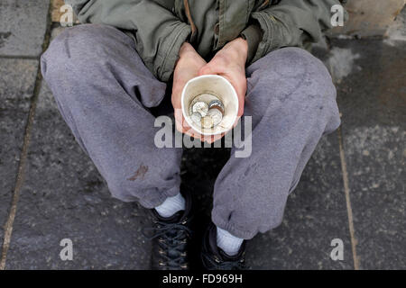 Street mendicante, seduto su un marciapiede con una tazza di cartone per la raccolta di fondi e di carità. Foto Stock