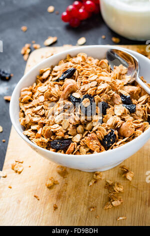 Muesli fatti in casa con i dadi e la frutta secca in bianco ciotola, un vasetto di yogurt fresco e frutti su sfondo di legno Foto Stock