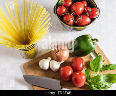 Ingredienti freschi della cucina italiana esposti su un tagliere di legno con coltello da chef, pasta di capelli d'angelo e un colino di ceramica di pomodori maturi di vite. Foto Stock