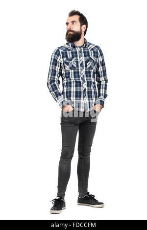 Grave barbuto hipster in controllato plaid shirt con le mani nelle tasche che guarda lontano. Corpo pieno lunghezza ritratto isolato su bianco Foto Stock