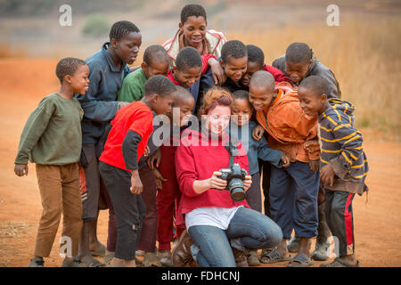 Giovani ragazzi africani in piedi intorno a una ragazza americana guardando la sua fotocamera nella regione Hhohho dello Swaziland, Africa. Foto Stock