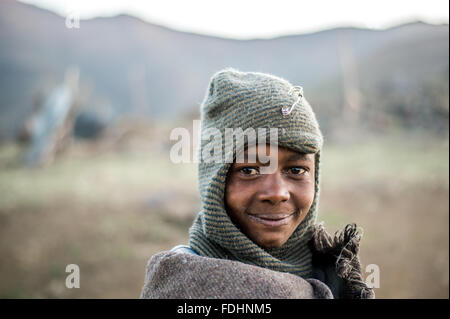 Ragazzo giovane avvolto in una coperta in piedi di fronte ad una gamma di montagna in Lesotho, Africa Foto Stock