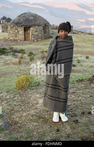 Ragazzo giovane avvolto in una coperta in piedi di fronte a una capanna di pietra in Lesotho, Africa Foto Stock