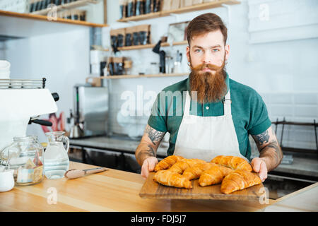 Attraente barbuto barista in piedi in cafe e di contenimento della scheda in legno con croissant Foto Stock