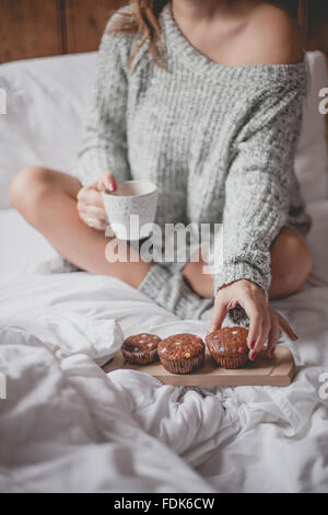 Donna seduta sul letto con la tazza di tè e caffè e muffin Foto Stock