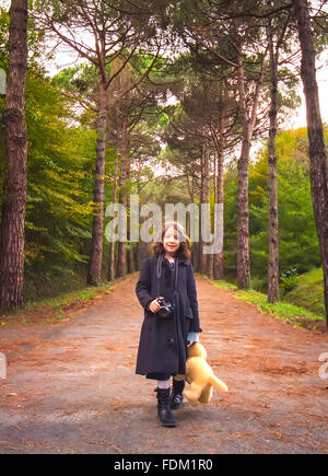 Bambina ha una macchina fotografica in mano e un orso giocattolo in un altro, in una giornata autunnale. Foto Stock