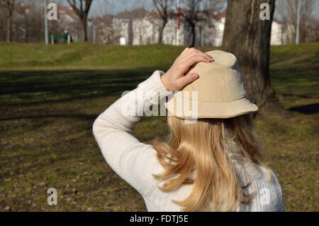 Donna con capelli biondi in un maglione bianco tenendo il suo cappello durante una passeggiata in un parco in una giornata di vento Foto Stock