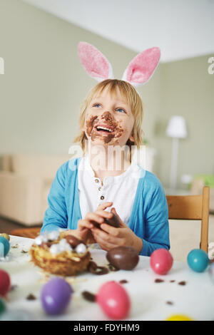 Felice ragazzo con orecchie di coniglio mangiando cioccolato uova da tavola di Pasqua Foto Stock