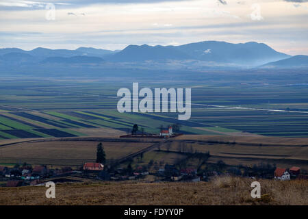 Piccolo villaggio con chiesa cattolica tra le colline e le montagne della Transilvania, la Romania in inverno senza neve Foto Stock